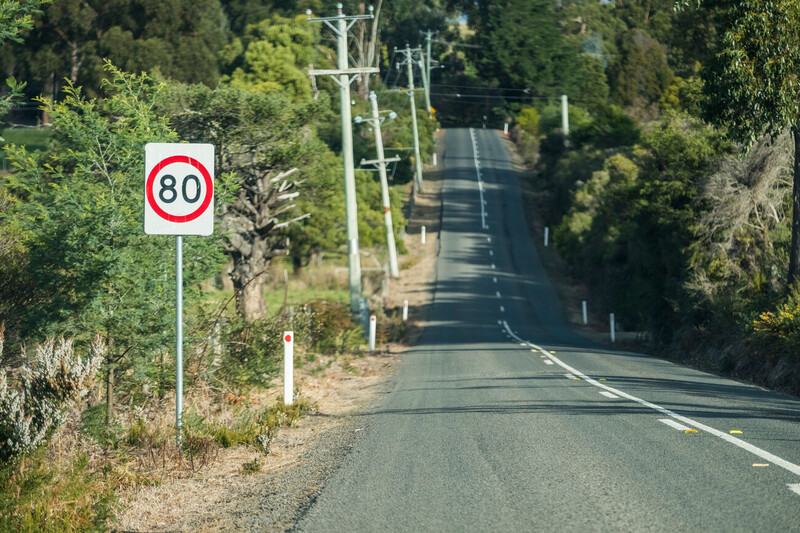 A sign displaying 80km speed limit on a sealed road lined with bushland