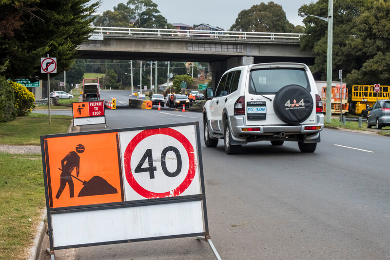 A sign displaying a 40km speed limit on a road with road work equipment including witches hats. A white SUV is driving past the sign.